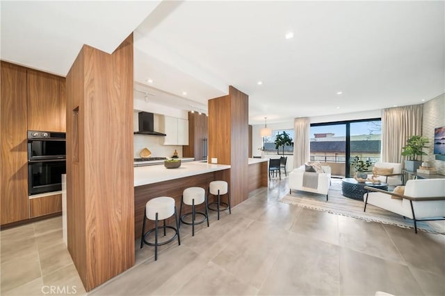 kitchen with dobule oven black, a kitchen breakfast bar, wall chimney exhaust hood, brown cabinetry, and modern cabinets