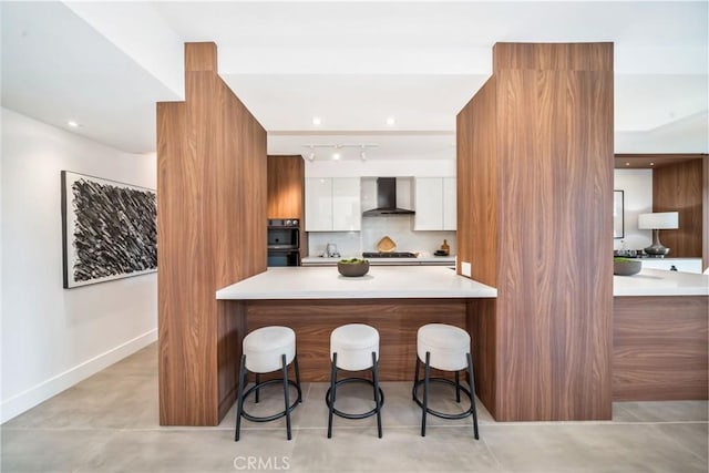 kitchen featuring a breakfast bar area, white cabinets, black double oven, gas cooktop, and wall chimney exhaust hood