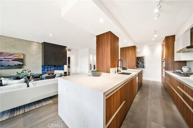 kitchen with an island with sink, sink, a large fireplace, stainless steel gas cooktop, and wall chimney exhaust hood