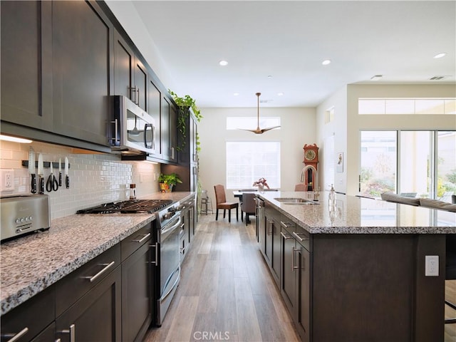 kitchen with sink, appliances with stainless steel finishes, a kitchen island with sink, light stone counters, and light hardwood / wood-style floors