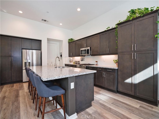 kitchen with sink, stainless steel appliances, light hardwood / wood-style floors, an island with sink, and light stone countertops