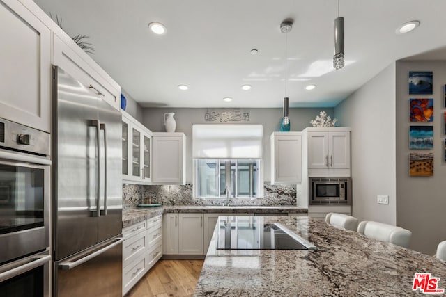 kitchen with hanging light fixtures, backsplash, built in appliances, light stone countertops, and white cabinets