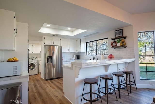 kitchen featuring white microwave, backsplash, stainless steel fridge with ice dispenser, a peninsula, and a raised ceiling