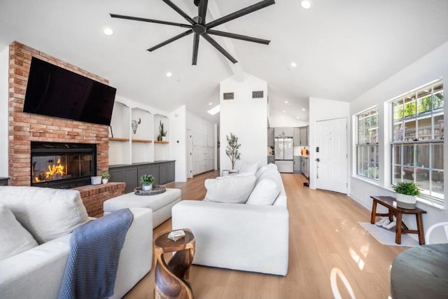 living room with ceiling fan, a brick fireplace, lofted ceiling with beams, and light hardwood / wood-style flooring