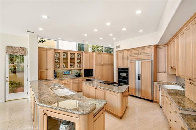 kitchen with light stone counters, light brown cabinetry, black appliances, and a center island with sink