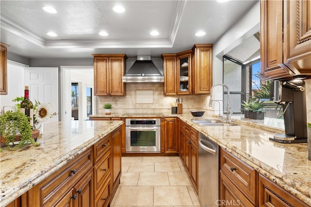 kitchen with wall chimney exhaust hood, sink, light stone counters, a raised ceiling, and stainless steel appliances