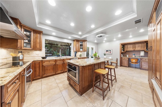 kitchen with wall chimney range hood, a center island, a raised ceiling, black electric cooktop, and oven