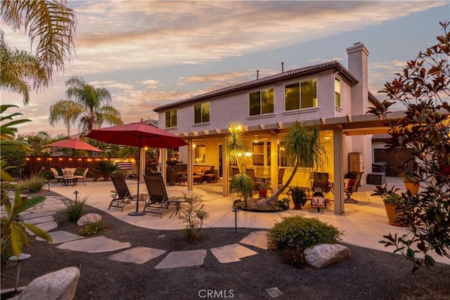 back of house at dusk with stucco siding, fence, a chimney, and a patio area