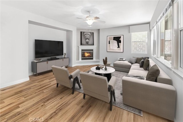 living room with a glass covered fireplace, baseboards, light wood-type flooring, and ceiling fan