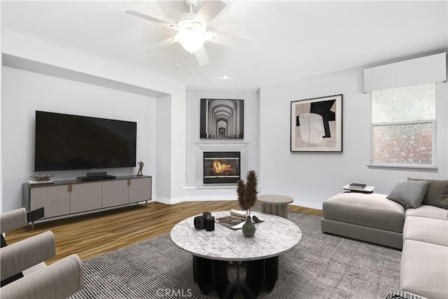 living area featuring baseboards, wood finished floors, a glass covered fireplace, and a ceiling fan