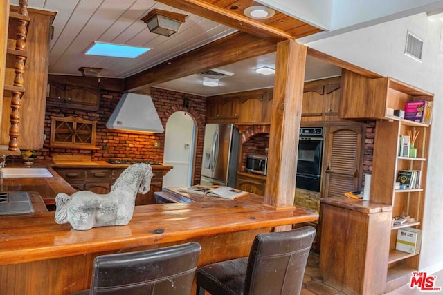 kitchen featuring beamed ceiling, sink, wooden counters, black appliances, and wall chimney range hood