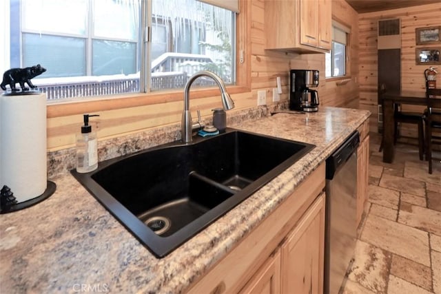 kitchen with light brown cabinetry, wooden walls, dishwasher, sink, and light stone countertops