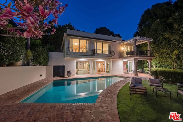 back house at night with a balcony, a yard, a patio area, and a pergola