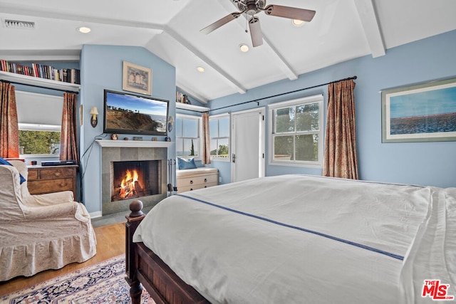 bedroom with hardwood / wood-style flooring, ceiling fan, a tiled fireplace, and lofted ceiling with beams