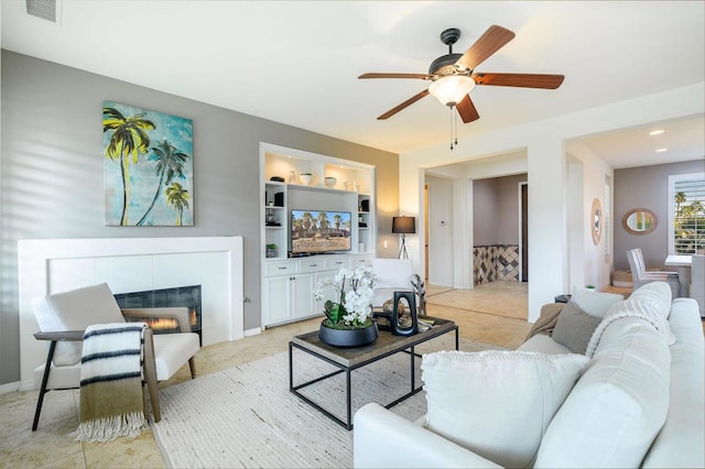 living room with a tiled fireplace, ceiling fan, and built in shelves