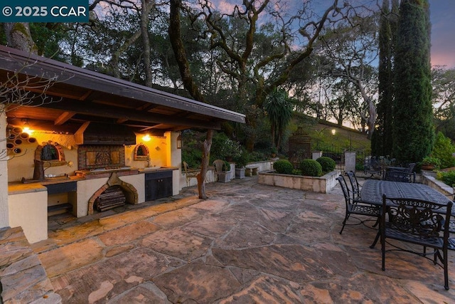 patio terrace at dusk with an outdoor fireplace and area for grilling