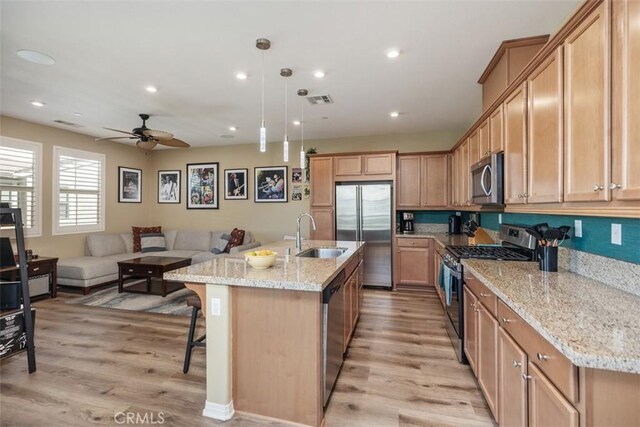 kitchen with sink, hanging light fixtures, stainless steel appliances, light stone countertops, and a center island with sink