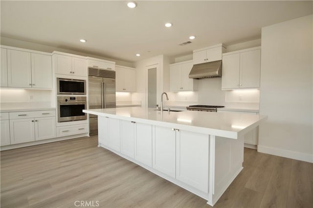 kitchen featuring light wood-style flooring, under cabinet range hood, a sink, white cabinets, and built in appliances