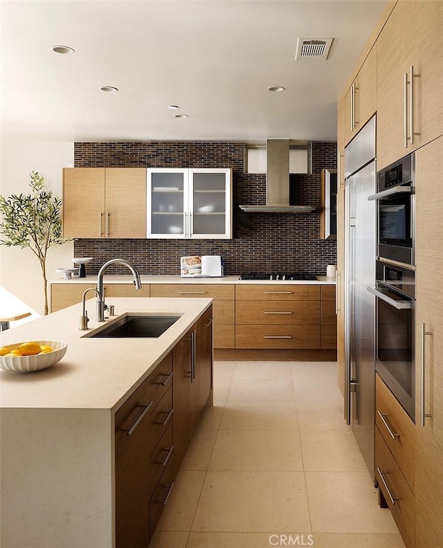 kitchen featuring light tile patterned flooring, wall chimney exhaust hood, sink, a kitchen island with sink, and backsplash