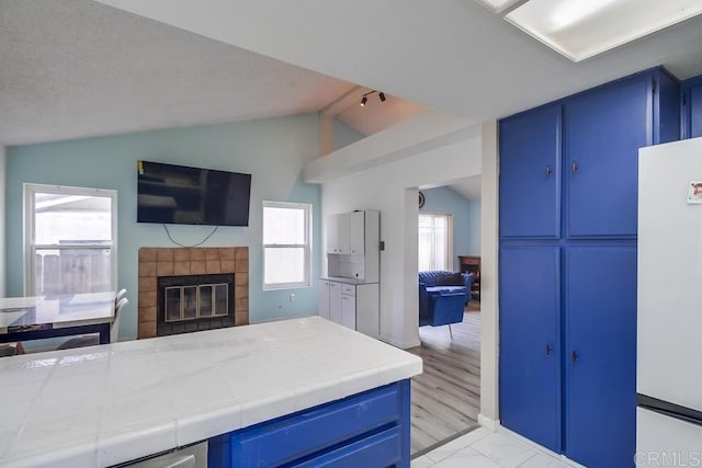 kitchen featuring vaulted ceiling, blue cabinets, a tiled fireplace, white refrigerator, and tile counters