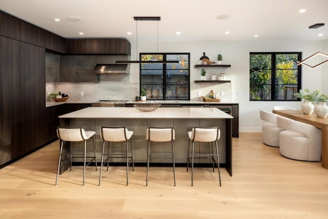 bar featuring dark brown cabinetry, tasteful backsplash, hanging light fixtures, light hardwood / wood-style flooring, and range hood