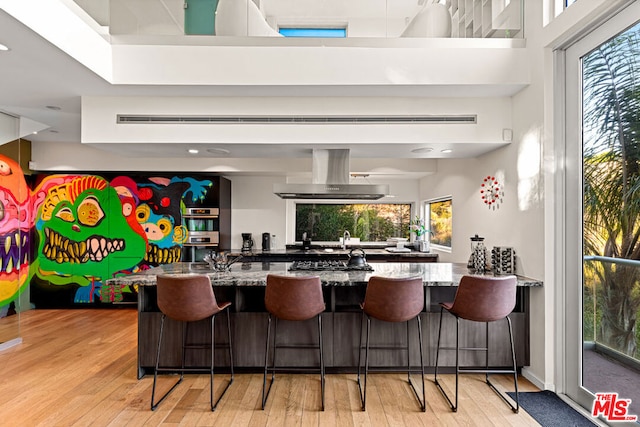 kitchen with a kitchen bar, sink, light hardwood / wood-style flooring, stone counters, and island exhaust hood