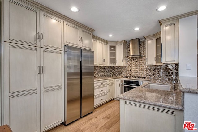 kitchen featuring wall chimney range hood, sink, appliances with stainless steel finishes, light hardwood / wood-style floors, and light stone countertops