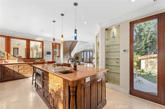 kitchen featuring a sink, a center island with sink, light stone countertops, stainless steel microwave, and decorative light fixtures