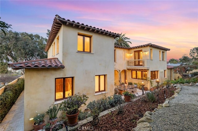 back of property at dusk with a balcony, a tiled roof, and stucco siding