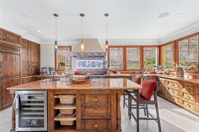 kitchen with wine cooler, light stone counters, ventilation hood, a center island with sink, and pendant lighting