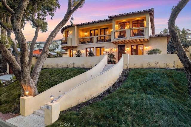 back of property at dusk with fence, a balcony, and stucco siding