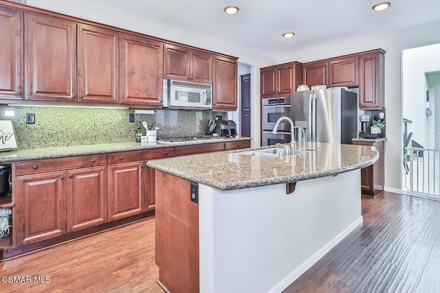 kitchen featuring sink, light stone counters, wood-type flooring, a center island with sink, and appliances with stainless steel finishes