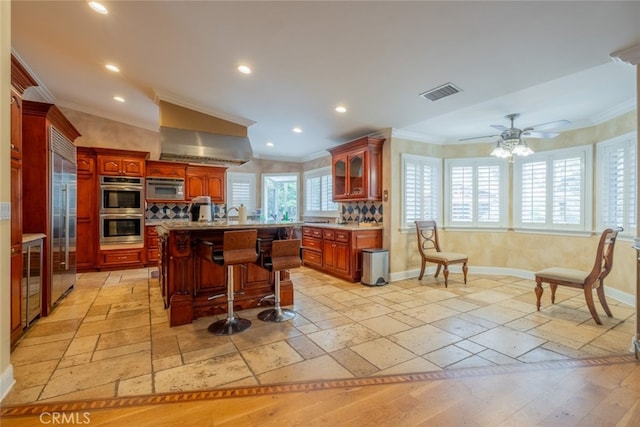 kitchen with a breakfast bar area, ceiling fan, built in appliances, ornamental molding, and an island with sink