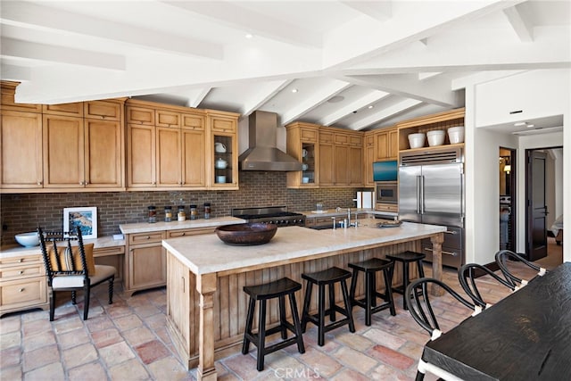 kitchen featuring vaulted ceiling with beams, built in appliances, tasteful backsplash, a center island with sink, and wall chimney exhaust hood