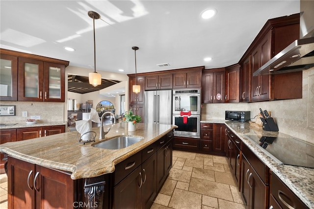 kitchen with pendant lighting, wall chimney range hood, sink, stainless steel appliances, and a center island with sink