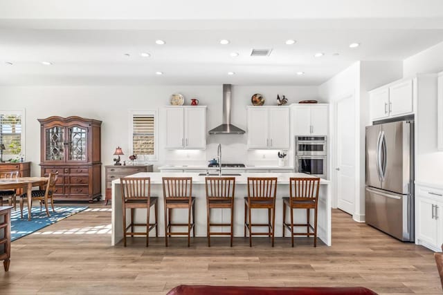 kitchen featuring appliances with stainless steel finishes, a breakfast bar, white cabinets, wall chimney range hood, and a center island with sink