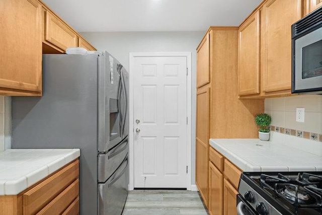 kitchen featuring light brown cabinetry, backsplash, tile counters, stainless steel appliances, and light wood-type flooring