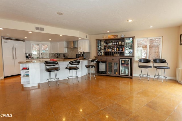 kitchen featuring backsplash, wine cooler, white cabinets, built in fridge, and wall chimney exhaust hood