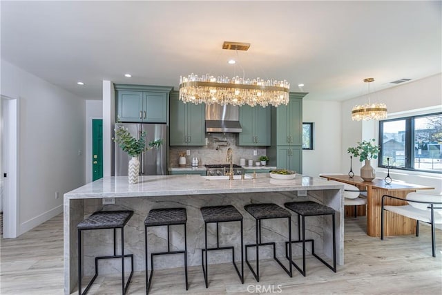 kitchen featuring a spacious island, wall chimney exhaust hood, light stone counters, hanging light fixtures, and stainless steel fridge