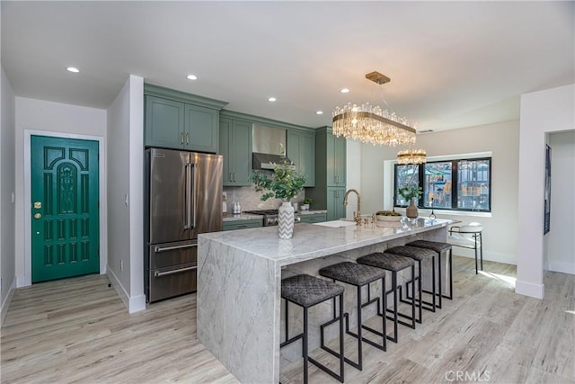 kitchen featuring high end refrigerator, decorative light fixtures, an island with sink, green cabinets, and light stone countertops