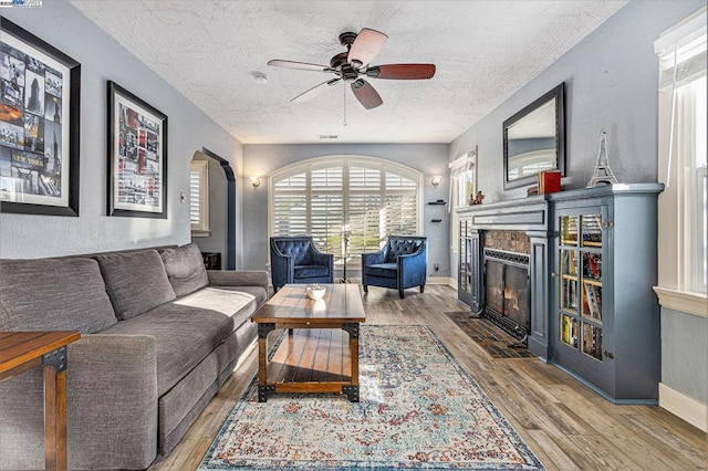 living room featuring ceiling fan, a stone fireplace, hardwood / wood-style floors, and a textured ceiling