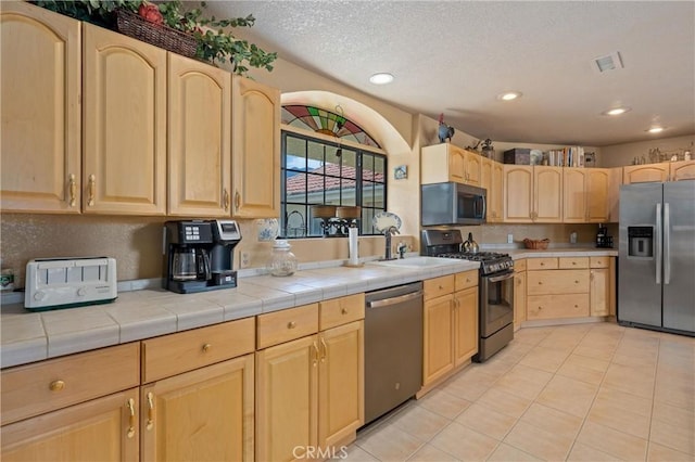 kitchen featuring tile countertops, light tile patterned floors, stainless steel appliances, light brown cabinets, and a textured ceiling