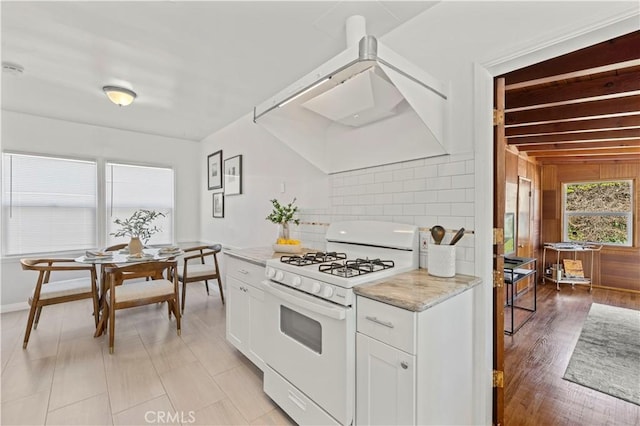 kitchen with white gas range oven, range hood, tasteful backsplash, light stone countertops, and white cabinets