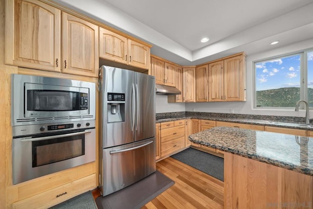 kitchen featuring appliances with stainless steel finishes, sink, dark stone countertops, light hardwood / wood-style floors, and light brown cabinets