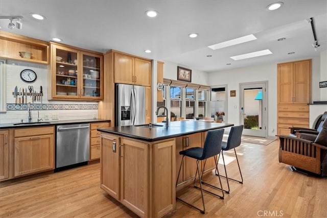 kitchen with a kitchen island with sink, stainless steel appliances, a sink, dark countertops, and glass insert cabinets