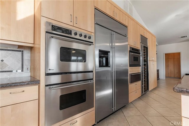 kitchen featuring light tile patterned flooring, dark stone countertops, decorative backsplash, stainless steel appliances, and light brown cabinets