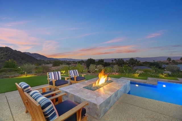 pool at dusk featuring a patio, a mountain view, a fire pit, and a lawn