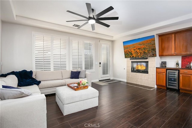 living room featuring ceiling fan, a tiled fireplace, bar area, dark hardwood / wood-style flooring, and beverage cooler