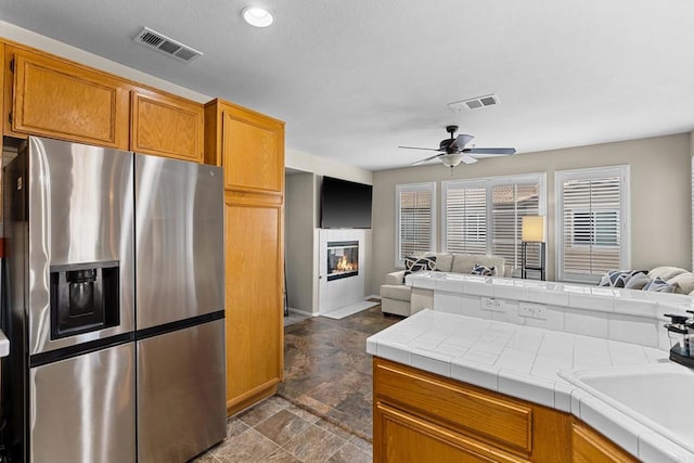 kitchen with sink, stainless steel fridge, ceiling fan, tile counters, and a tiled fireplace