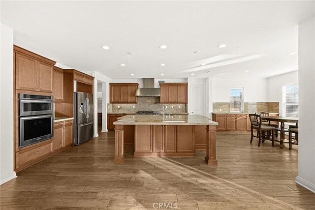 kitchen featuring wood-type flooring, an island with sink, wall chimney exhaust hood, and appliances with stainless steel finishes